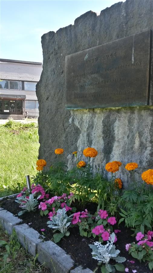 Kivessä on metallinen muistolaatta. Muistomerkin edessä on kukkaistutuksia.