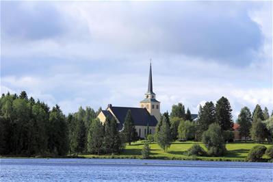 Vaalean kellertevä kirkkorakennus Kiurujärven rannalla. Kellotorni kohoaa korkeana kirkon päädyssä. Kirkkoa ympäröivät suuret koivut sekä rannan salavat ja kuuset.