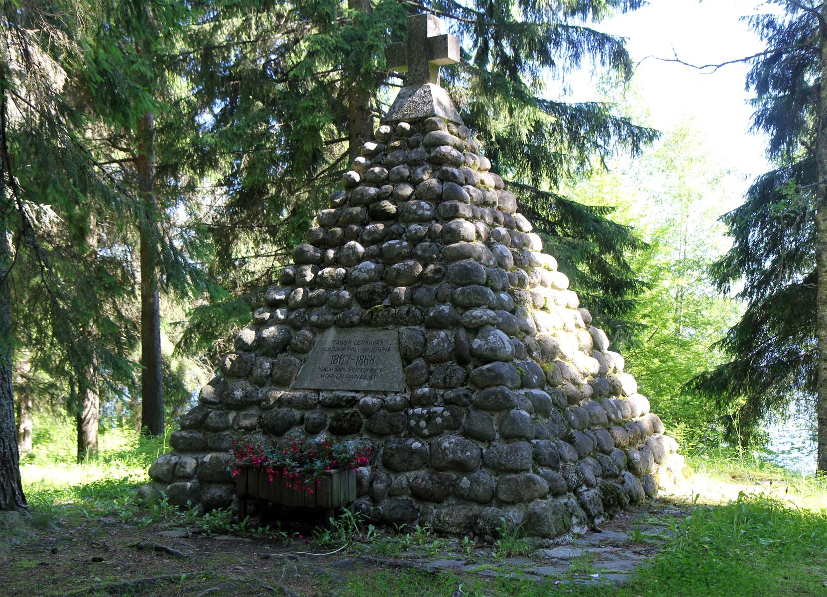 Pyramidin muotoinen kivimuodostelma puiden katveessa. Pyramidin huipulla on risti ja kyljessä muistolaatta.